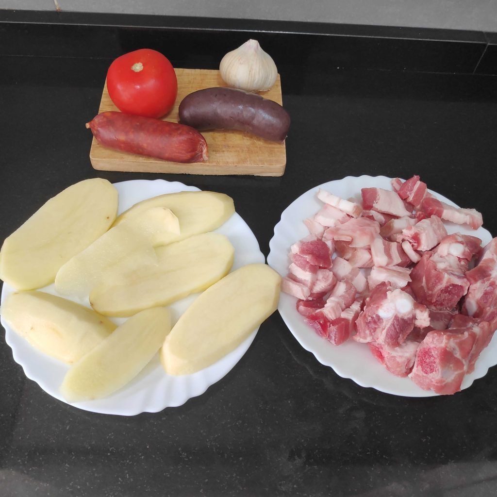 dos platos blancos con patata uno y el otro con costilla y panceta a trozos, al fondo una madera de cortar con tomate, una cabeza de ajos y un chorizo y una morcilla