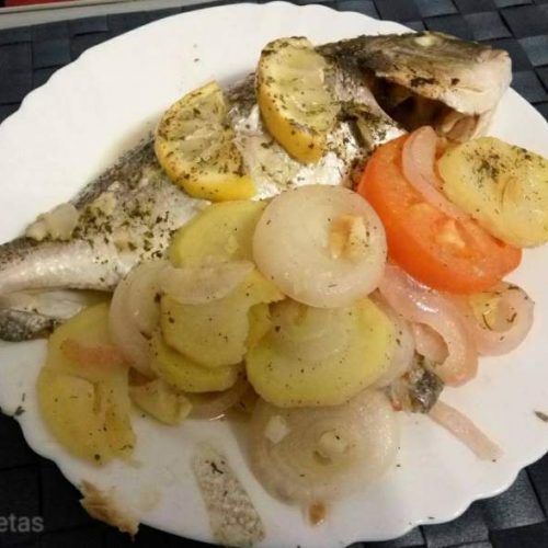una orada al forn amb patates en un plat blanc amb ceba, i tomàquet, porta rodanxes de llimona