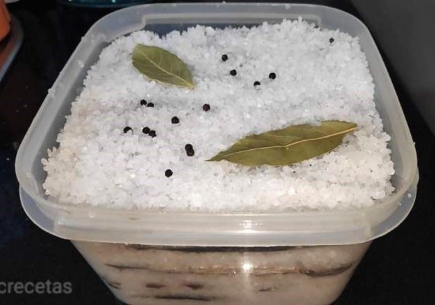 recipiente lleno de boquerones y sal gorda, arriba granos de pimienta negra y una hoja de laurel