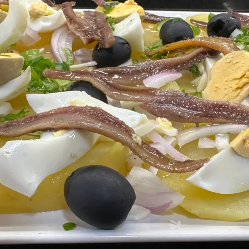 una ensalada de patatas y anchoas con huevo duro y olivas negras en una fuente blanca rectangular